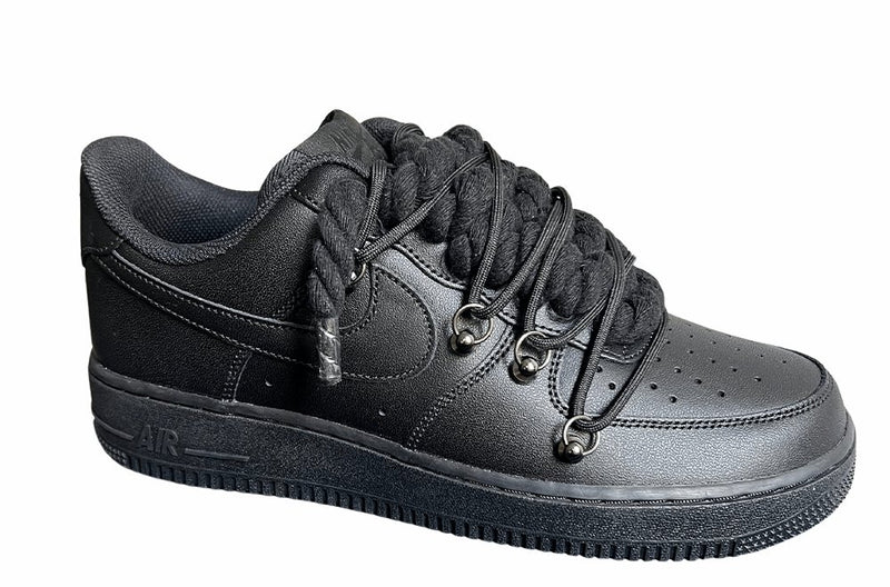 Schwarzer Nike Air Force 1 Sneaker mit dicken schwarzen Rope-Schnürsenkeln, kombiniert mit schwarzen Akzent-Schnüren. Der Schuh hat eine matte schwarze Oberfläche, Metallösen und eine robuste Sohle, die dem Design einen modernen und markanten Look verleiht, von der Seite.