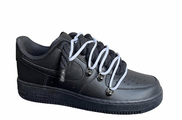 Schwarzer Nike Air Force 1 Sneaker mit dicken schwarzen Rope-Schnürsenkeln, kombiniert mit weißen Akzent-Schnüren. Der Schuh hat eine matte schwarze Oberfläche, Metallösen und eine robuste Sohle, die dem Design einen modernen und markanten Look verleiht, von der Seite.