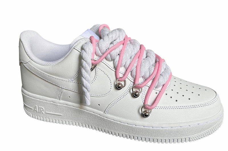 Weißer Nike Air Force 1 Sneaker mit dicken weißen Rope-Schnürsenkeln, kombiniert mit pinken Akzent-Schnüren. Der Schuh hat eine matte weiße Oberfläche, Metallösen und eine robuste Sohle, die dem Design einen modernen und markanten Look verleiht, von der Seite.