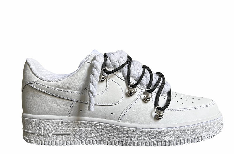 Weißer Nike Air Force 1 Sneaker mit dicken weißen Rope-Schnürsenkeln, kombiniert mit schwarzen Akzent-Schnüren. Der Schuh hat eine matte weiße Oberfläche, Metallösen und eine robuste Sohle, die dem Design einen modernen und markanten Look verleiht.